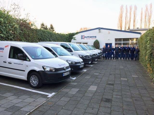 Mehrere weiße Firmenfahrzeuge mit „Kaufmanns“-Logo parken vor dem Firmengebäude, daneben steht das Team in blauer Arbeitskleidung.