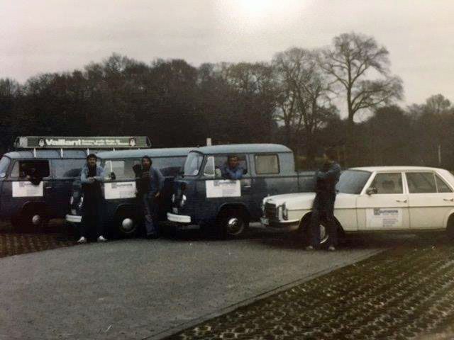 Historisches Foto mit drei alten Fahrzeugen und fünf Personen, aufgenommen auf einem Parkplatz vor einer Baumreihe.
